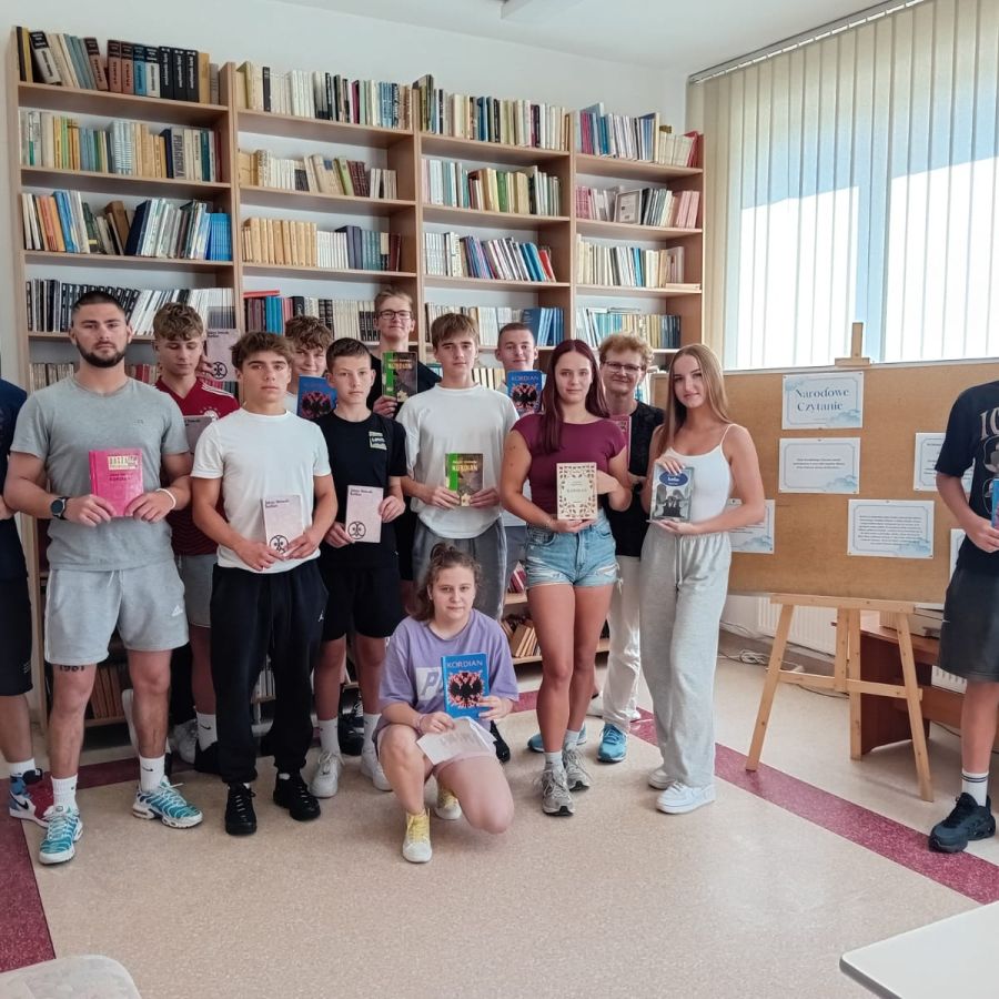 Uczniowie czytający Kordiana stoją w szkolne bibliotece. Po prawej stronie opis wydarzenia, w tle - regały z książkami.